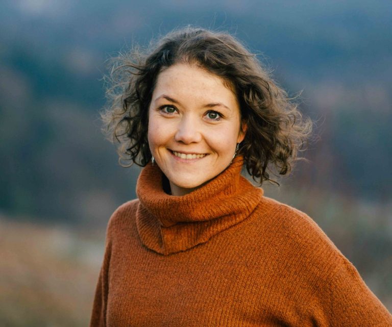 Deine Fotografin mit Herz Fotografin mit lockigem Haar, lächelnd in orangefarbener Rolle steht vor verschwommenem Hintergrund.